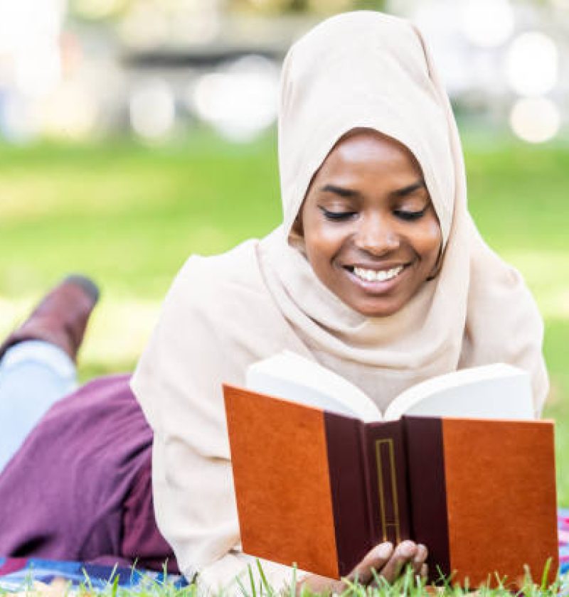 Muslim beautiful black woman wearing a hijab lying on the floor at a public park reading a book